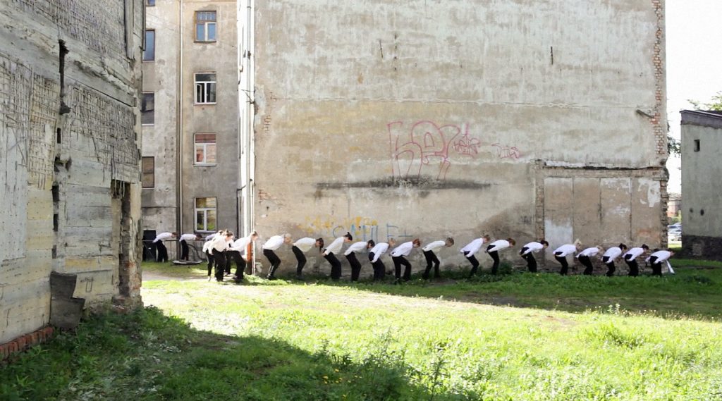 People bent over, standing in a meadow in front of a wall