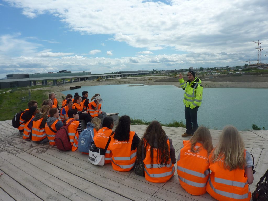 Schüler*innengruppe mit orangen Jacken am See sitzend