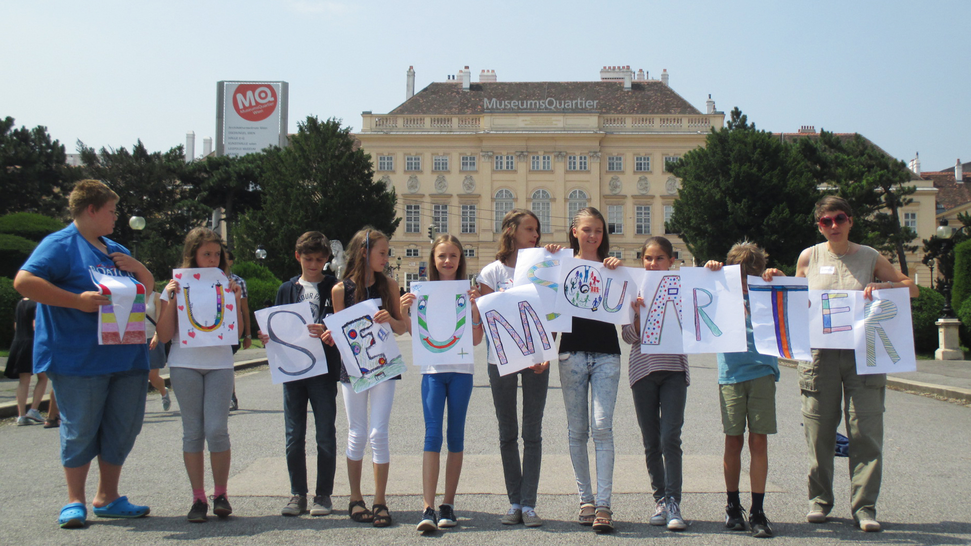 10 Kinder stehen vor dem Museumsquartier und halten gemalte Buchstaben hoch, die das Wort Museumsquartier ergeben