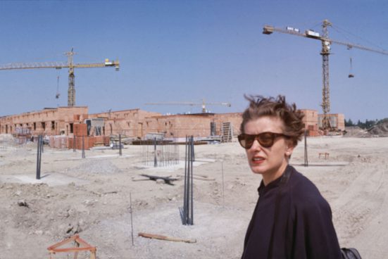 Eine Frau mit Sonnenbrille und kurzen Haaren und schwarzer Bluse auf einer Baustelle stehend, dahinter zwei Kräne und blauer Himmel