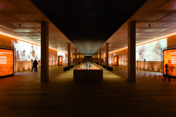 Interior view of an elongated exhibition space with columns and a long display case in the middle and projections on the side walls