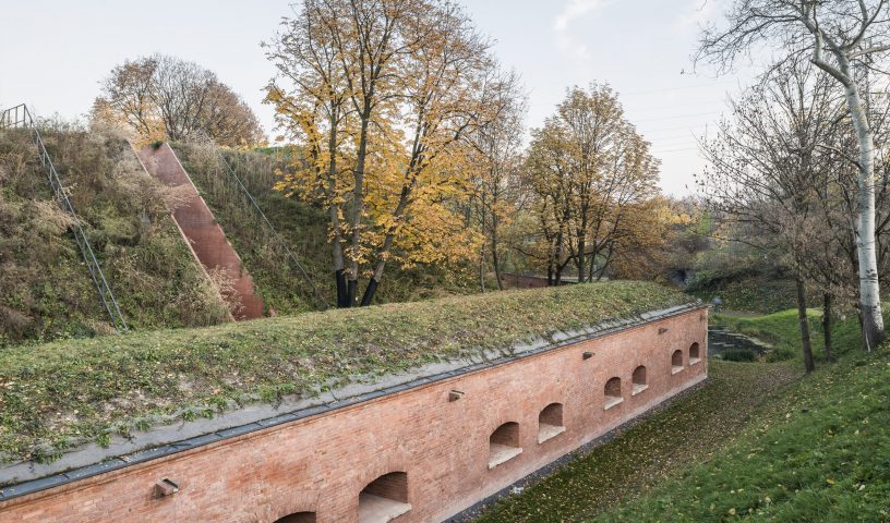 Wall with small window openings, grass-covered roof, surrounded by green space