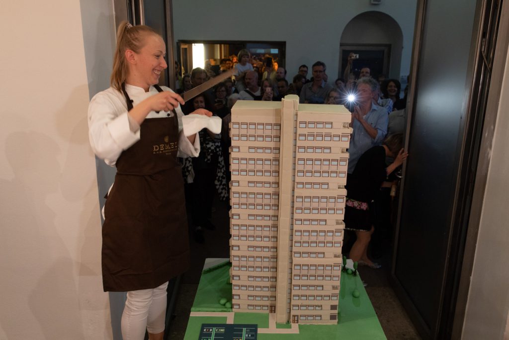 eine Tortenbäckerin mit großem Messer und einer Schürze schneidet einen Kuchen in vorm eines Hochhauses an