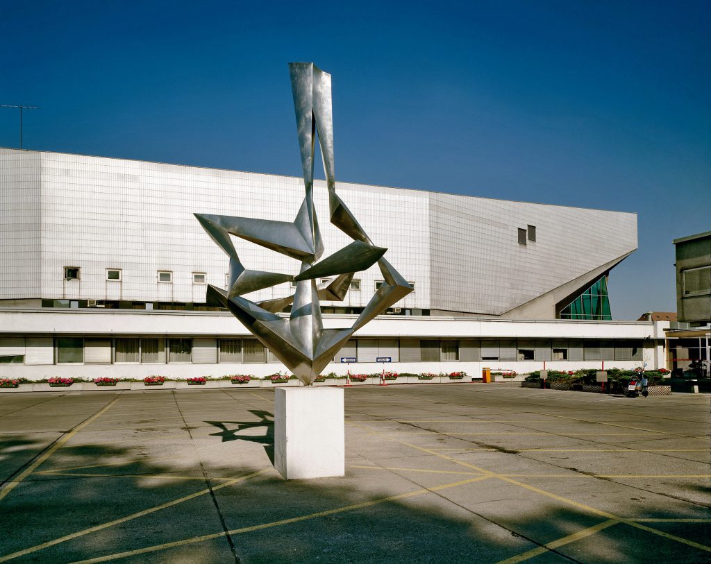 Exterior view of a hall with a concrete facade; in the foreground a square with a modern sculpture