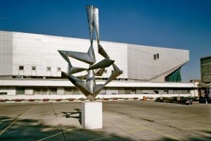Exterior view of a hall with a concrete facade; in the foreground a square with a modern sculpture