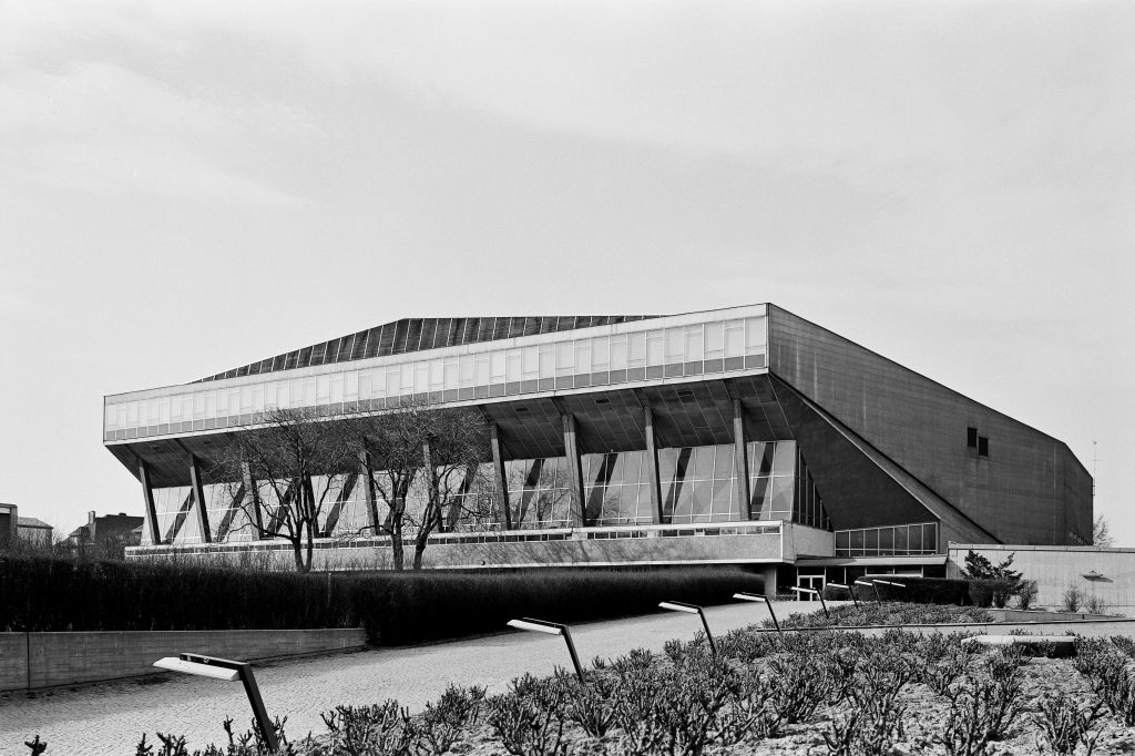 Schwarz-weiß Außenansicht eines Hallenbaus. Die Fassade ist mit Säulen versehen. Im Vordergrund eine Straße mit Straßenlaternen und eine Grüngrünfläche