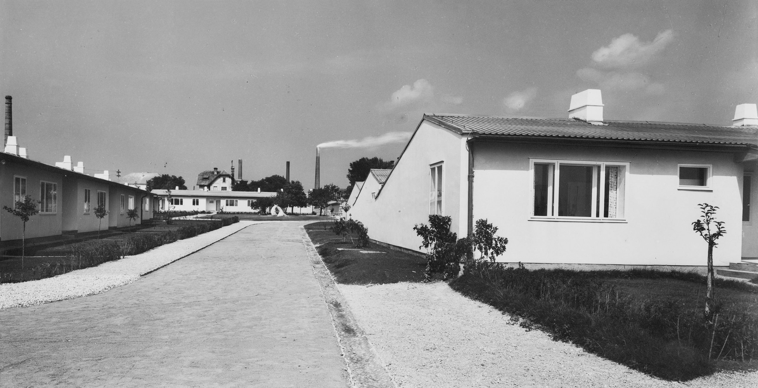 schwarz-weiß Ansicht eines Straßenzugs mit kleinen eingeschoßigen Wohnhäusern mit Satteldach. Fabriksschlote im Hintergrund