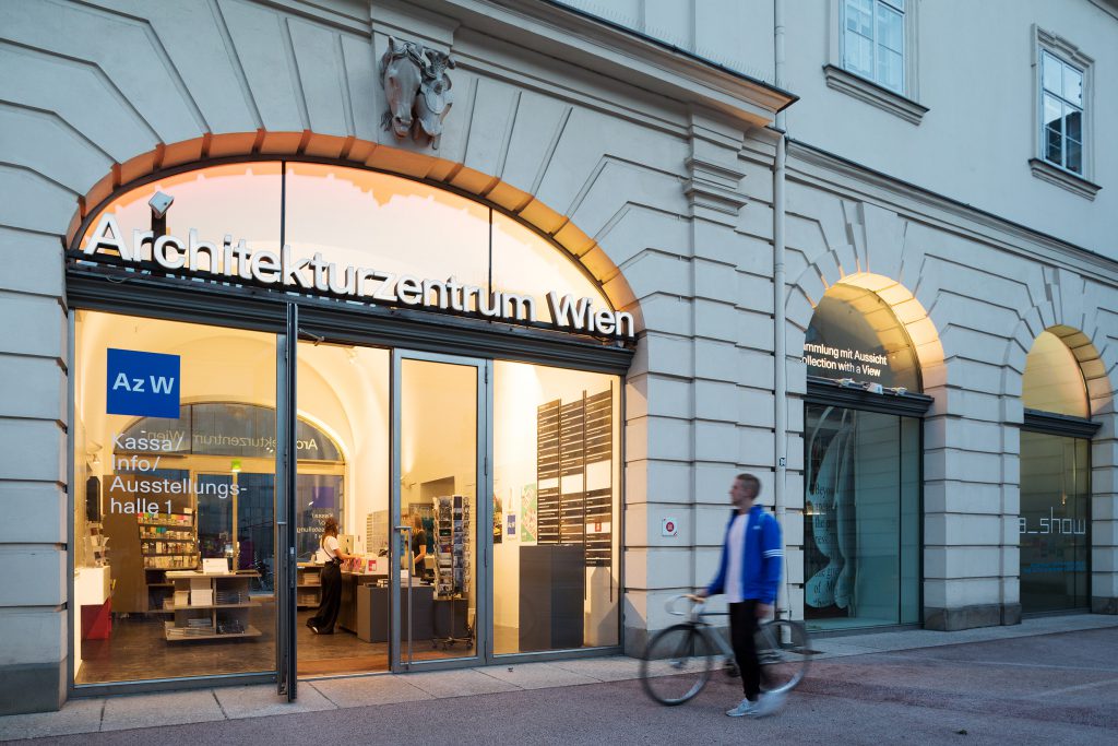 Mann mit blauem Annorak schiebt sein fahrrad am erleuchteten Eingangsbereich des Az W vorbei