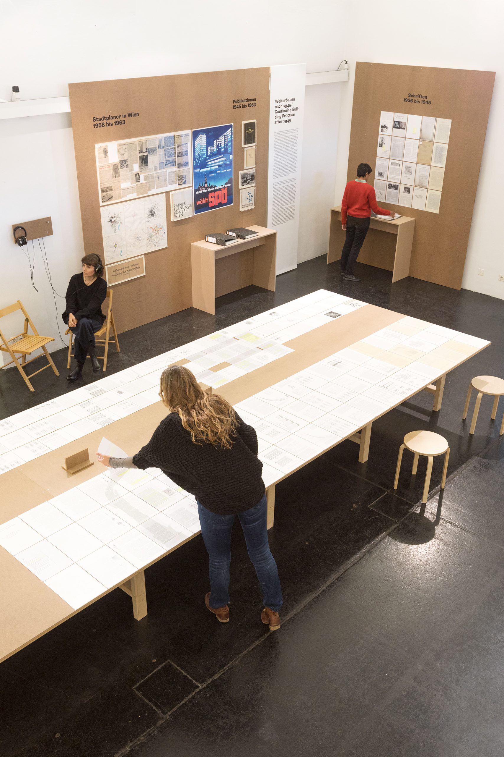 Drei Frauen in einem Ausstellungsraum. Eine Frau sitzt und trägt Kopfhörer. Die beiden anderen stehen und lesen in den ausgestellten Unterlagen.