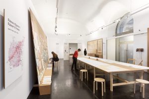 Three women in an exhibition room with a large table in the middle. One woman sits and reaches for a document, the other two stand and look at the exhibited documents or plans.