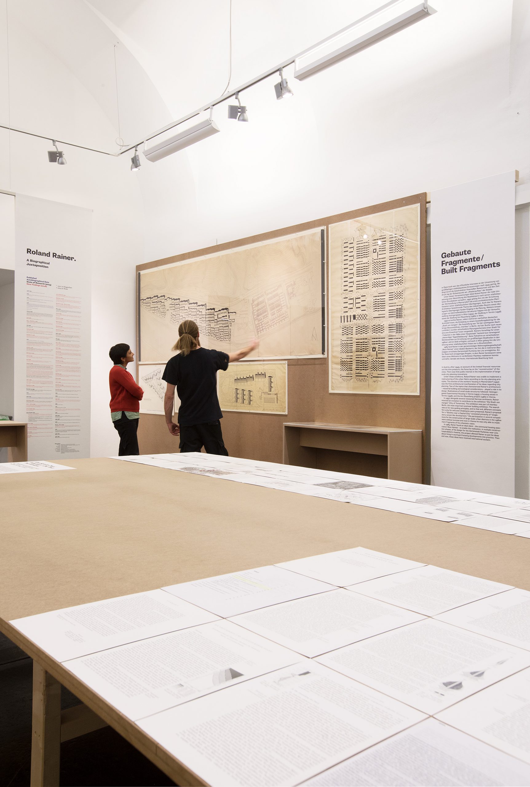 Ein Mann und eine Frau betrachten einen Plan, der an einer Ausstellungswand hängt. Der Mann deutet mit der Hand auf den Plan. Ein großer Tisch mit ausgestellten Dokumenten steht im Vordergrund