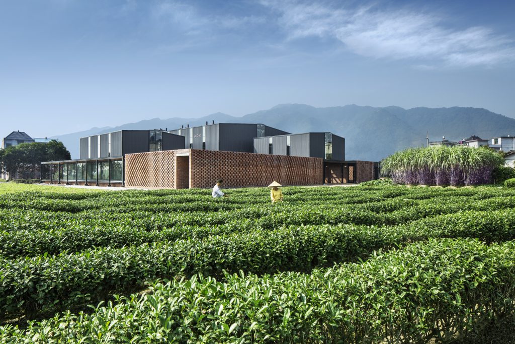 Cube-shaped, brown flat building with three elevated grey elements; in the foreground a tea plantation with 2 workers