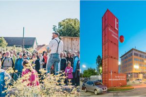 Ein Mann steht erhöht und sprich zu einer Gruppe von Menschen; rechts ein Turm aus roten Containern