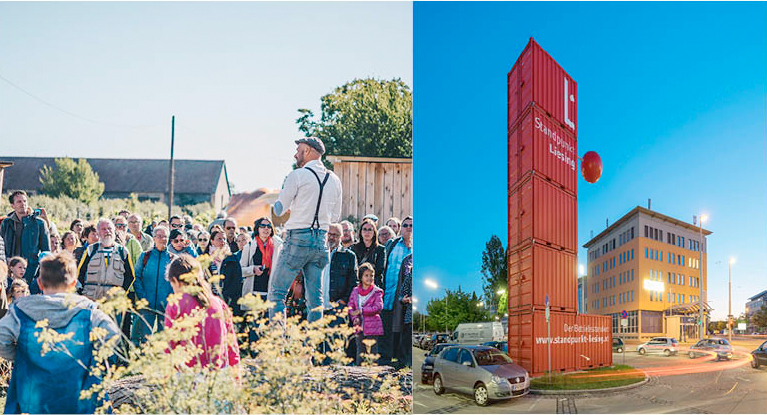 A man stands elevated and speaks to a group of people; on the right a tower made of red containers