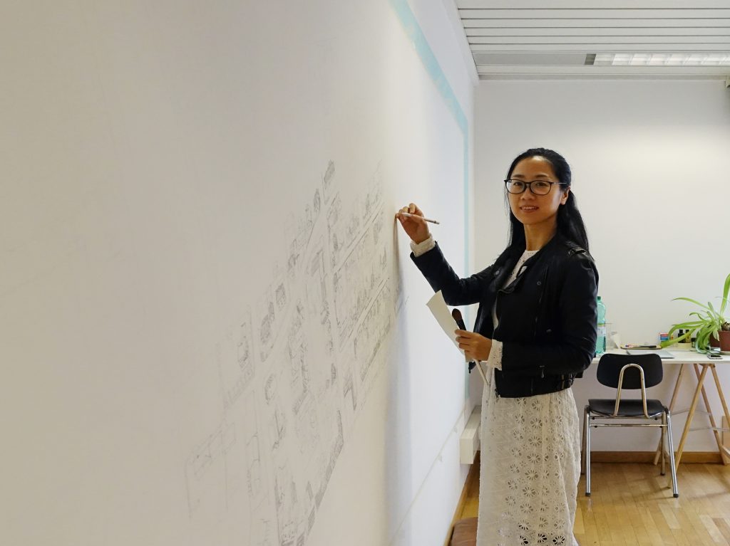 A woman with glasses draws a village on a white wall.