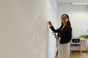 A woman with glasses draws a village on a white wall.