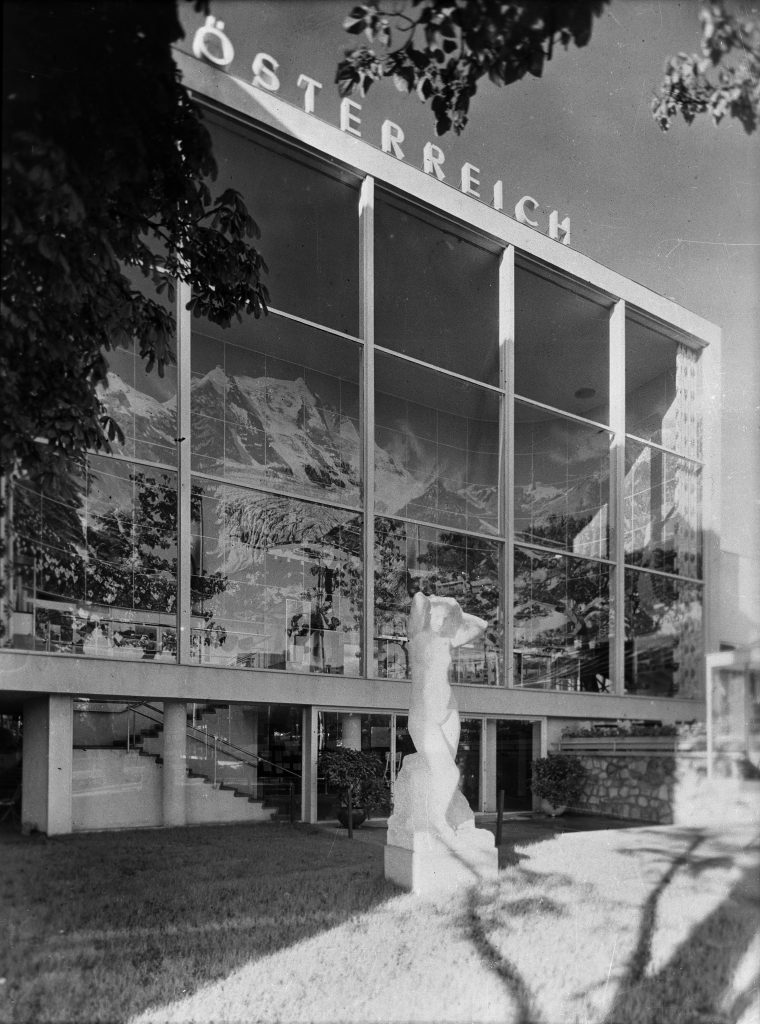 schwarz-weiß Bild einer Glasfassade, dahinter Aufnahmen eines Bergpanoramas, davor weiße Frauenskulptur