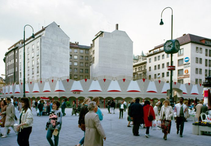 gemusterter farbiger Zaun mit hohen Gebäuden dahinter und Passant*innen im Vordergrund