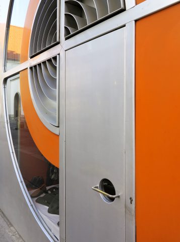 metallene Türe, daneben ein halbrundes Schaufenster und orange Wand