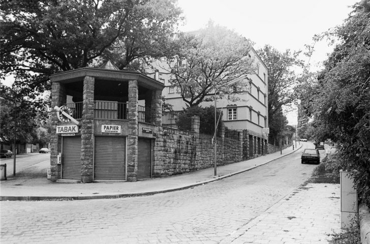 Schwarz-weiß-Foto eines mehrstöckigen Hauses und eines Pavillons neben einer Straße mit geparkten Autos