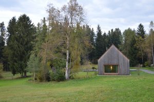 Holzhaus mit eine großen Fenster und satteldach auf einer Wiese neben einem Baum und Wald im Hintergrund