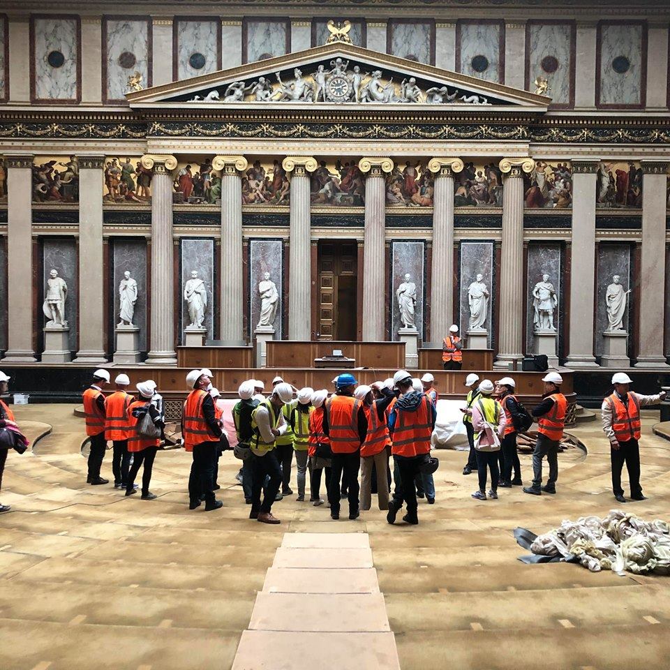 Gruppe von Menschen mit weißen Helmen und Warnwesten in historischem Saal mit griechischen Säulen und Figuren