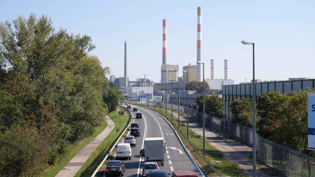 Autos auf einer mehrspurigen Straße und Fabriksschlote im Hintergrund