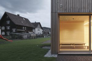 Section of a window with a view into the illuminated interior with a table and benches. Next to it there is an older wooden house.