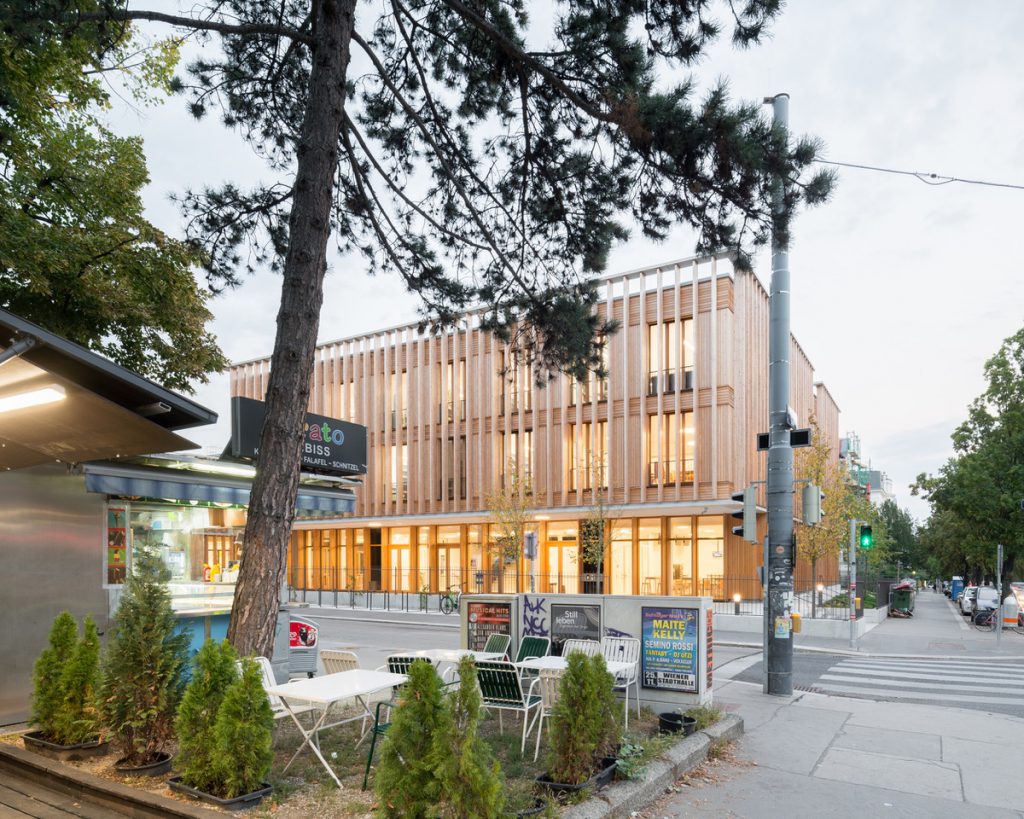 wooden building with lights in the groundfloor and trees in the foreground