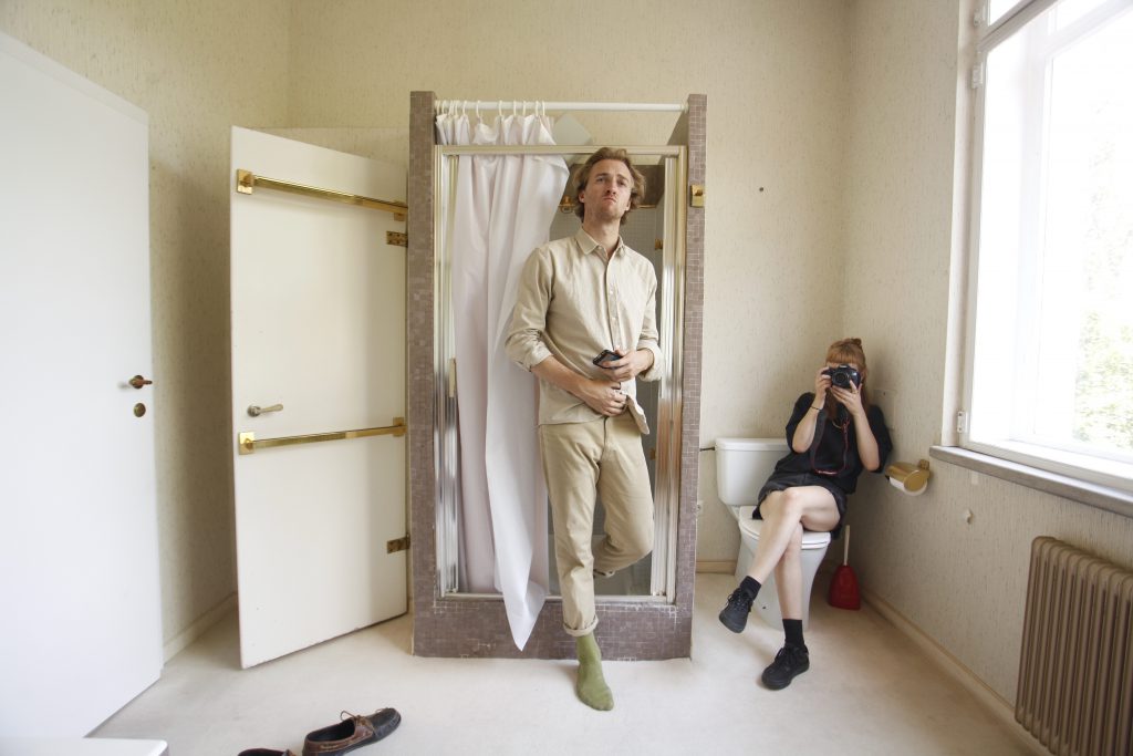 dressed man in shower, woman sitting beside with camera