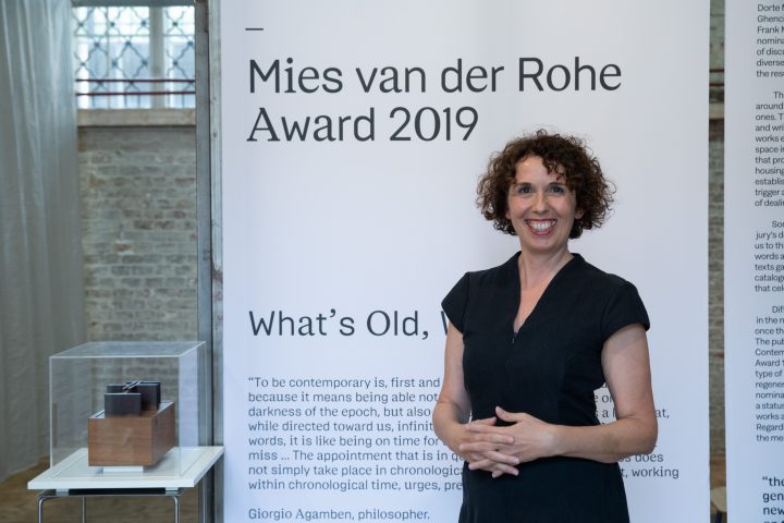 Woman with dark curly hair and black dress in front of white wall