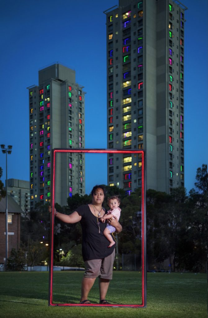 Eine Frau mit einem Kind am Arm auf einer Wiese vor Hochhäusern, einen roten Rahmen haltend
