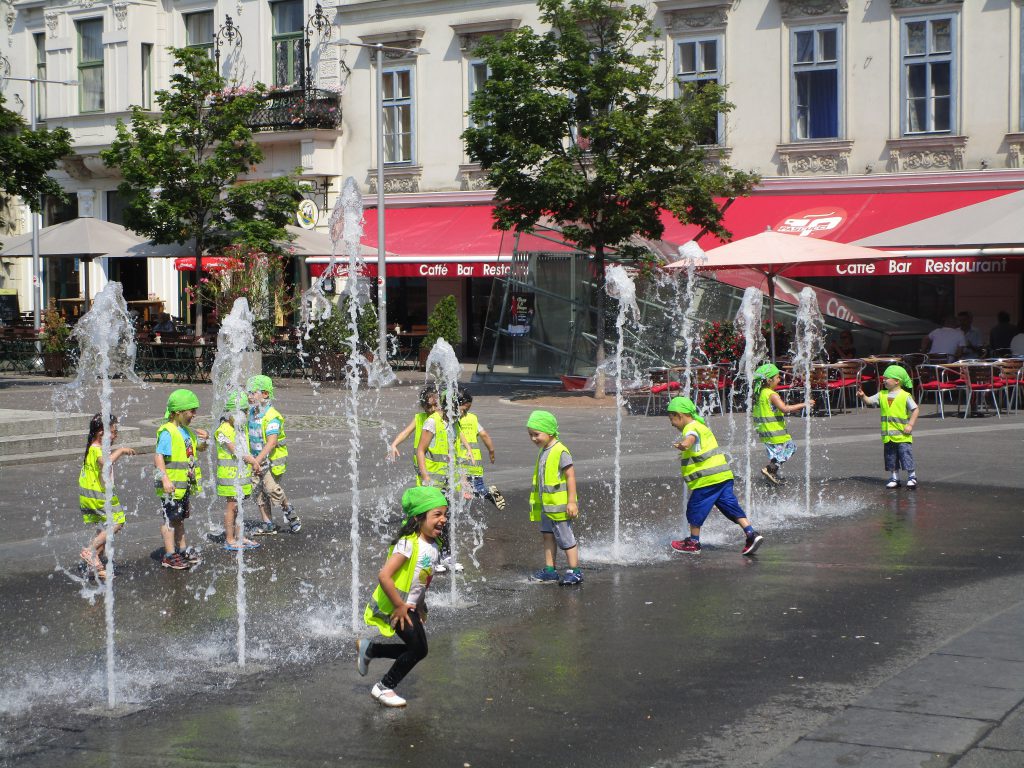 Kinder in Schutzwesten auf einem Platz mit Wasserbespielung