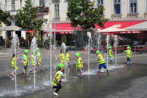 Kinder in Schutzwesten auf einem Platz mit Wasserbespielung