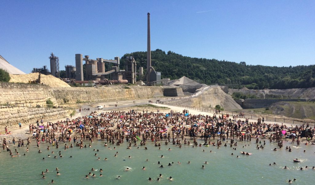 many people in and around a lake, behind it a factory