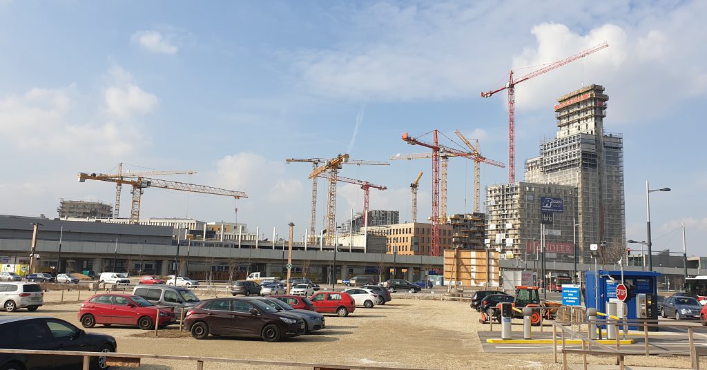 Zehn Baukräne vor einer Baustelle mit großem Parkplatz