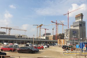 Ten construction cranes in front of a construction site with a large parking lot
