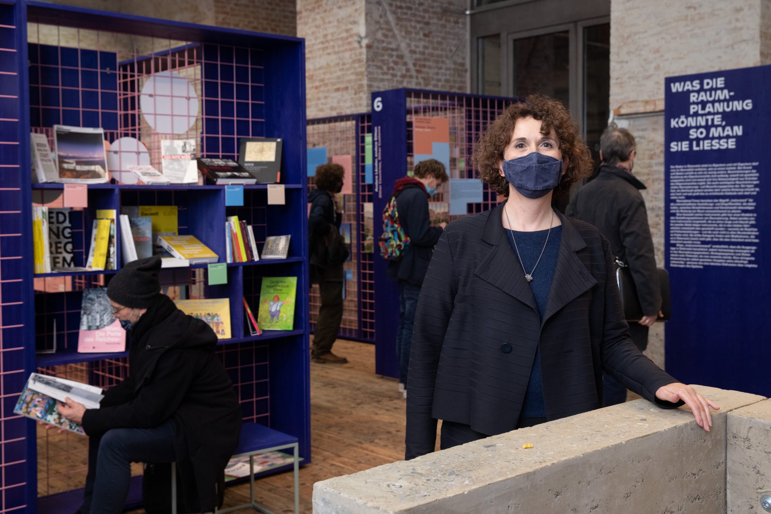 Frau mit dunklen Locken steht bei Betonwand in einer Ausstellung