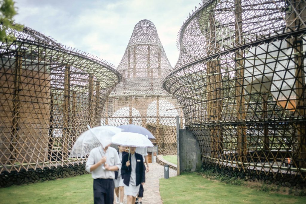 People with white umbrellas walk between bamboo buildings