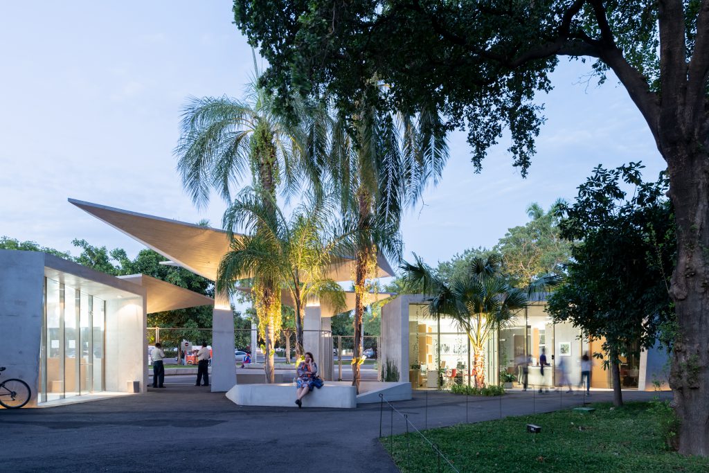 Entrance area at dusk with palm trees and trees, in front of it a woman sits on a white stone bench