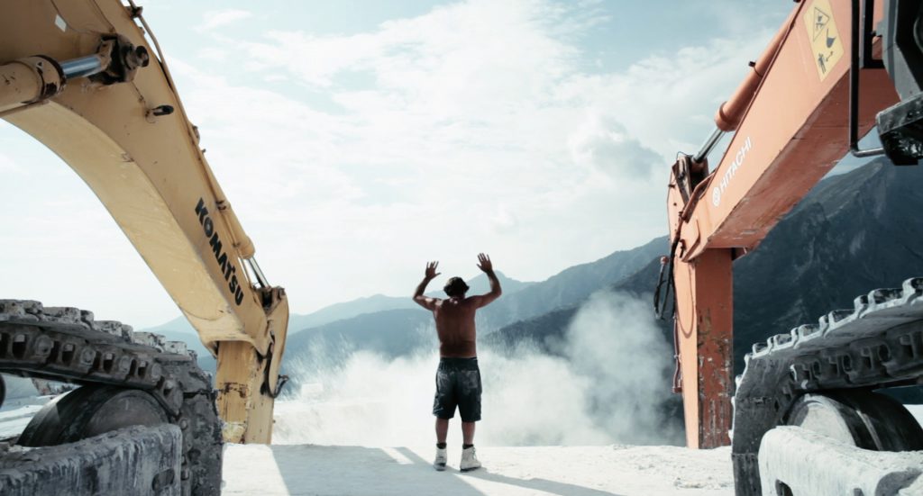 Man with raised arms between 2 large excavators