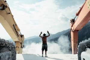 Man with raised arms between 2 large excavators