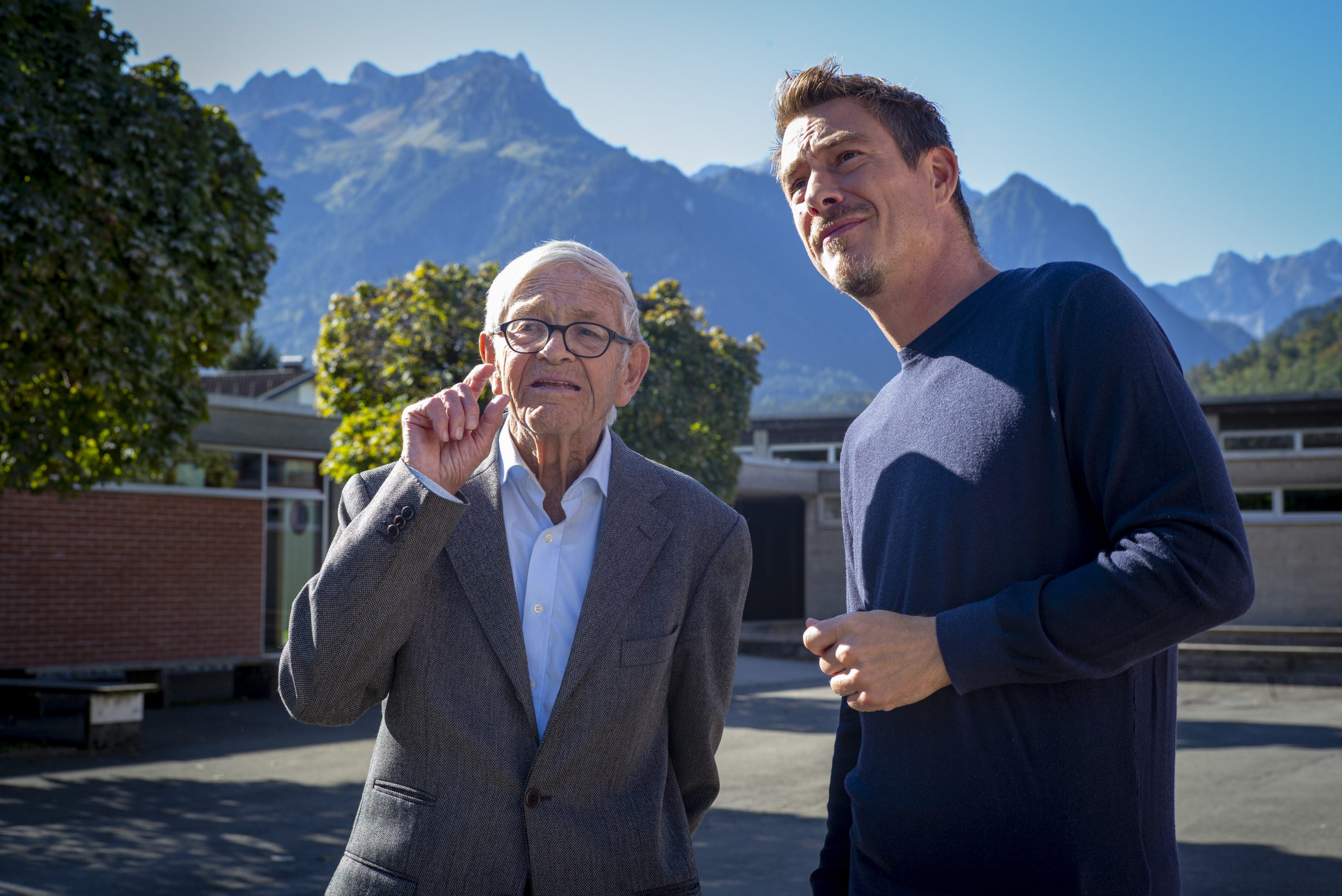 an old man with glasses and a young man stand in the sunshine in front of a mountain backdrop