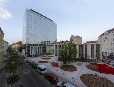 High building with glass façade in front of square with circular floor elements on which plants grow