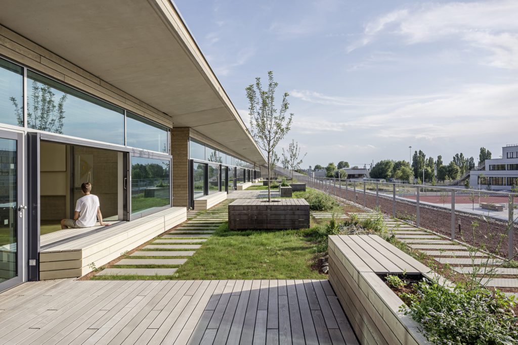 Blick auf eine große Terrasse mit Holzböden, Holzbänken und eine Fensterfront, eine Person sitzt auf einer Holzbank