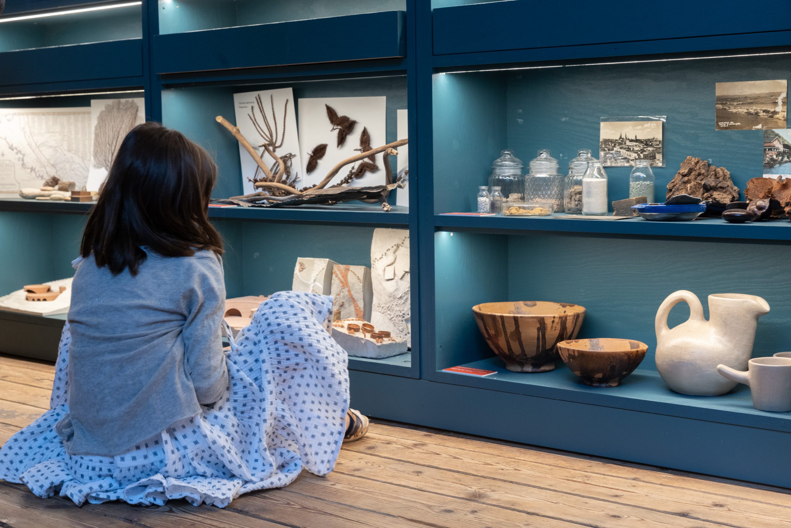 Girl in dress sits in front of wall with various objects