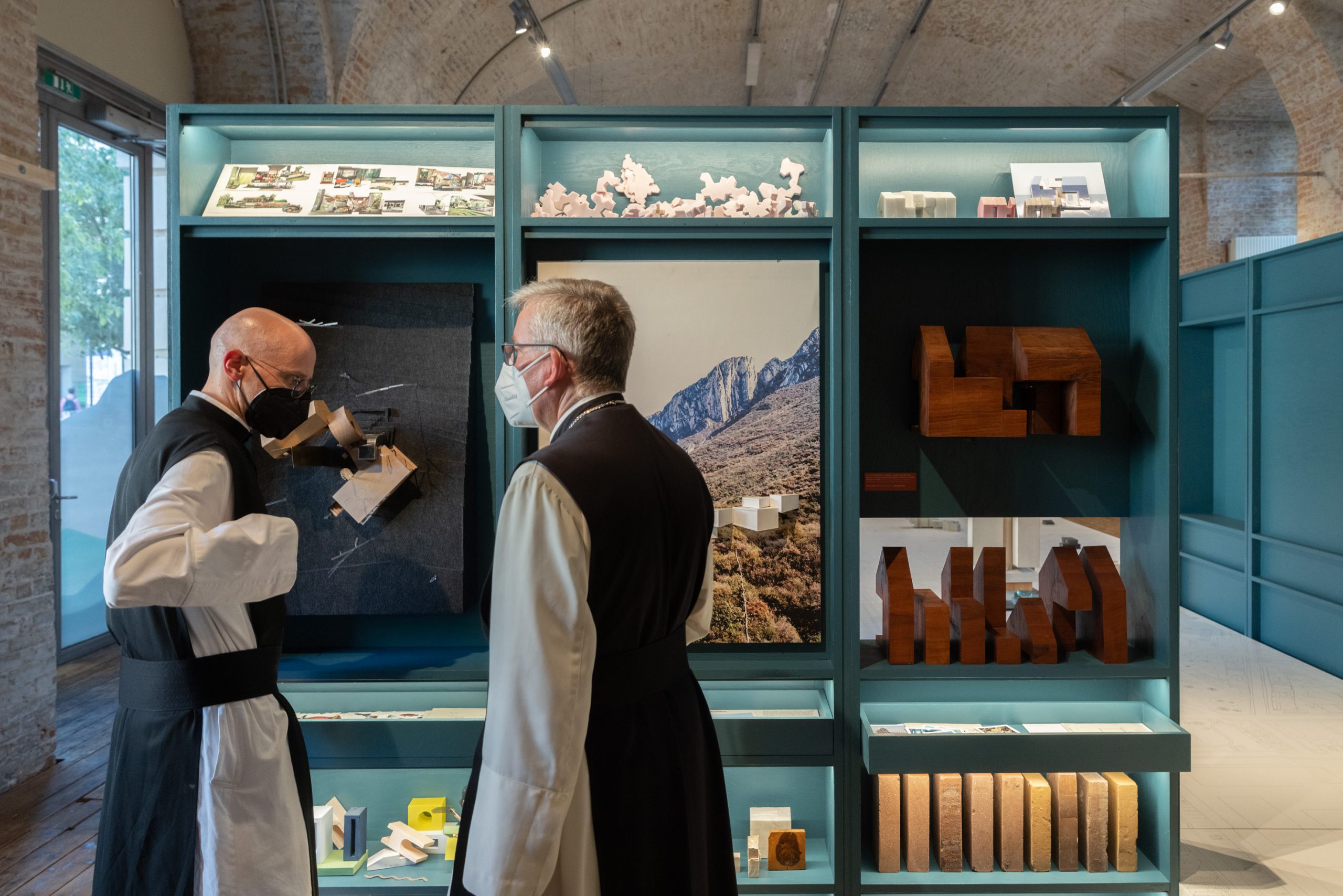 2 monks standing in front of a wall of shelves with various objects