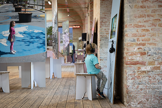 A child in a turquoise outfit sits with headphones in front of a wall, behind him large picture panels hanging from the ceiling.