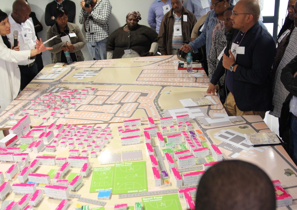 A model city on a large table. People stand and sit around it and discuss with each other