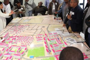 A model city on a large table. People stand and sit around it and discuss with each other